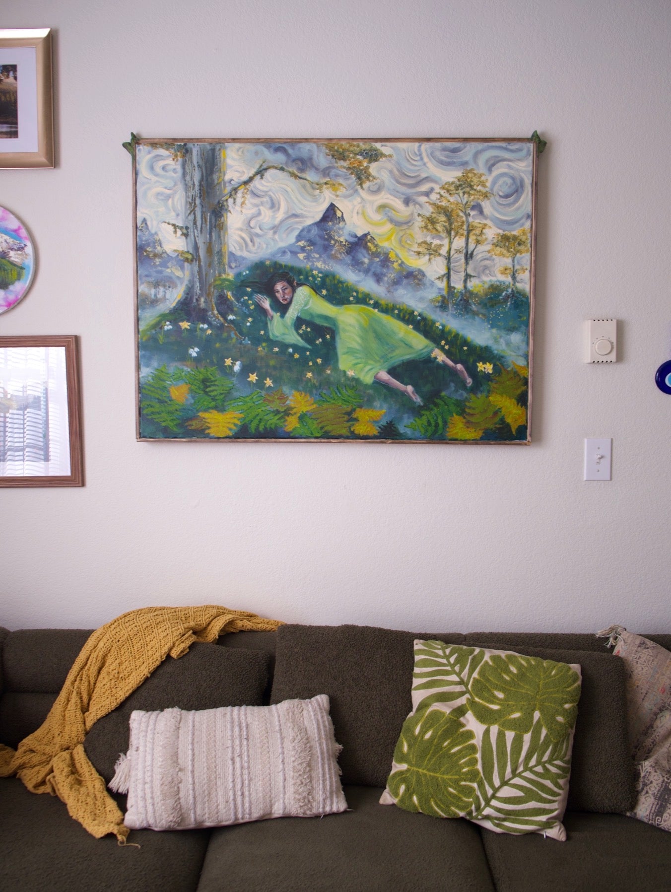 A framed art print depicting a woman in a green dress lying in a forest landscape, hanging on a light gray wall above a brown sofa with decorative pillows and a yellow throw blanket.
Arwen's Sorrow, Original Oil Painting with Custom Frame - Inspired by Lord of the Rings. Painting by Kelsey Weirick